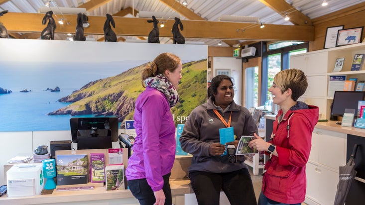Two visitors chatting to a member of staff with reception desk behind and photo of the coastline